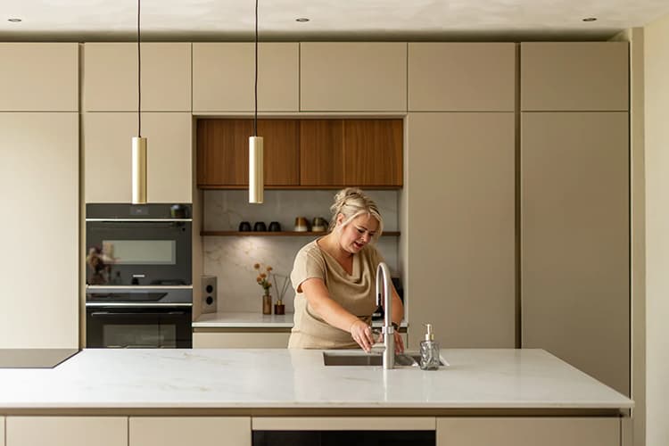 teaser-binnenkijken-bij-karina-en-roel.jpg Beige keuken met kookeiland waar een vrouw een glas water uit de kraan pakt